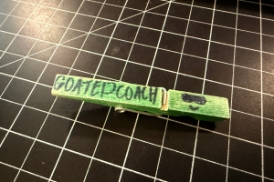 Half of a clothespin colored in green with the phrase "GOATED COACH" handwritten in big block letters.