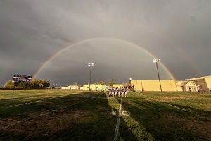 Football players on a field under a double rainbow a few moments before kickoff