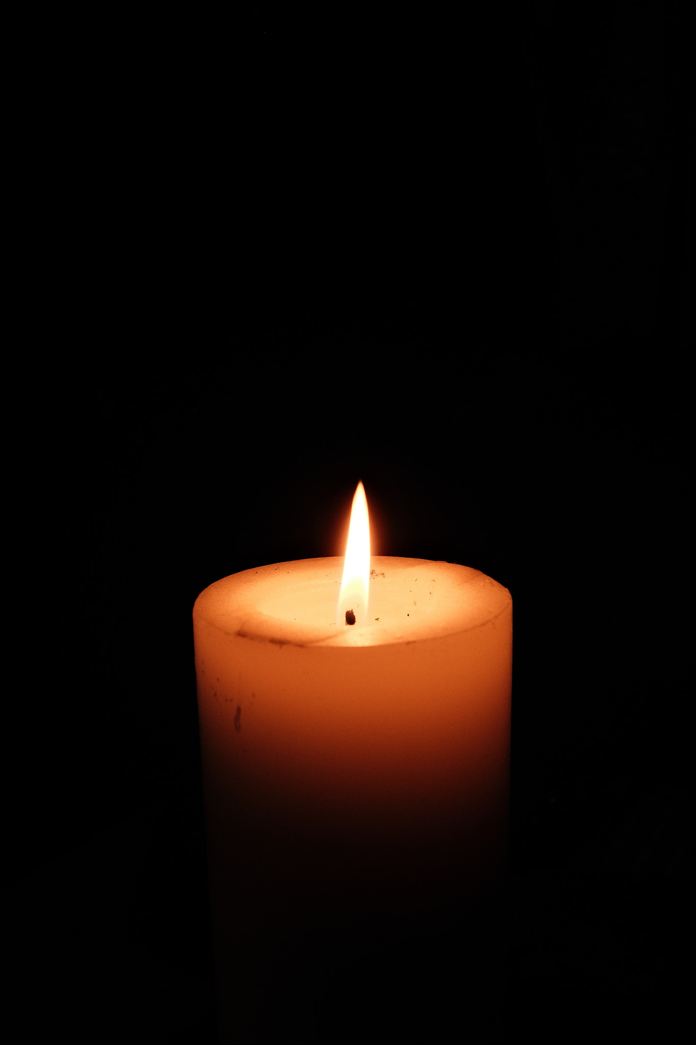 A single white pillar candle burning against a completely black background, its flame casting a warm light